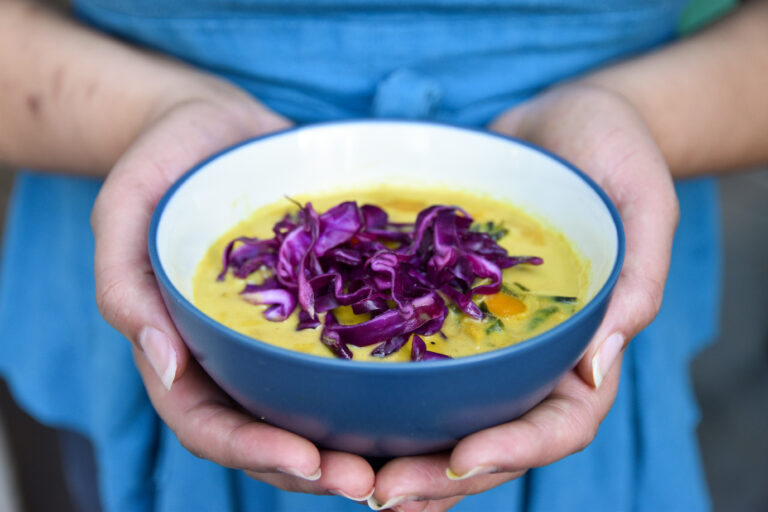 Two hands hold a bowl of yellow winter squash curry, topped with pickled purple cabbage pieces, served in a navy blue bowl.