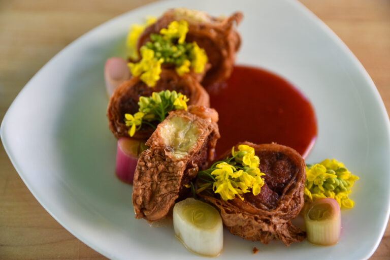 Pork kikiam (ground meat wrapped in tofu skin), garnished with yellow broccoli flowers, and served on top of red sauce, plated on a white ceramic dish. At the Foodwise Classroom at the Ferry Plaza Farmers Market in San Francisco.