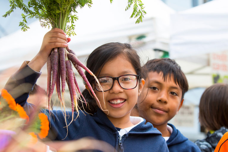 A girl holding a bunch of carrots next to a boy in the Ferry Plaza Farmers Market